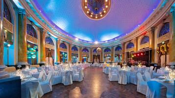 A grand ballroom with ornate columns, chandeliers, and tables set for a formal event.