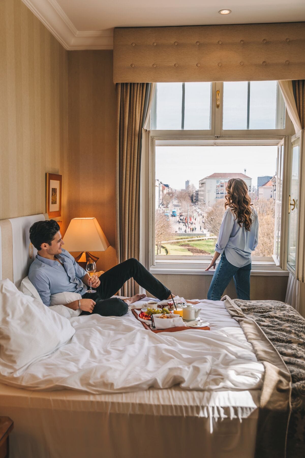 Et hotelværelse med en seng, et vindue med udsigt over byen, en lampe og et bakke med morgenmad.