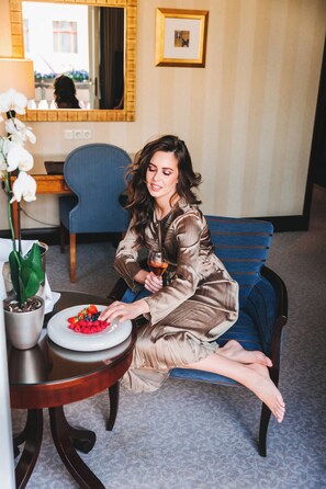 A woman sitting on a blue chair in a room with a potted plant, a table with a plate of fruit, and a mirror on the wall.