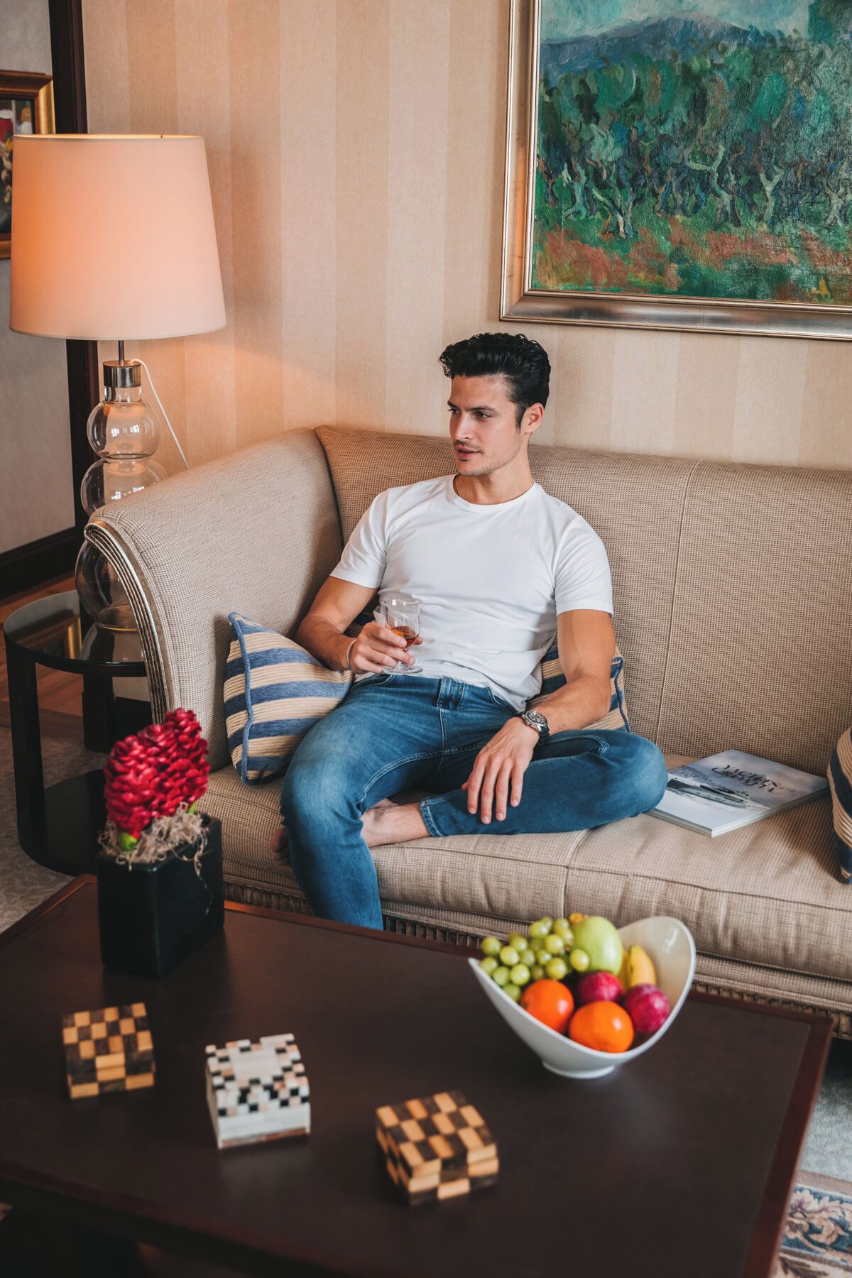 Un homme est assis sur un canapé beige, avec une lampe et un tableau en arrière-plan.