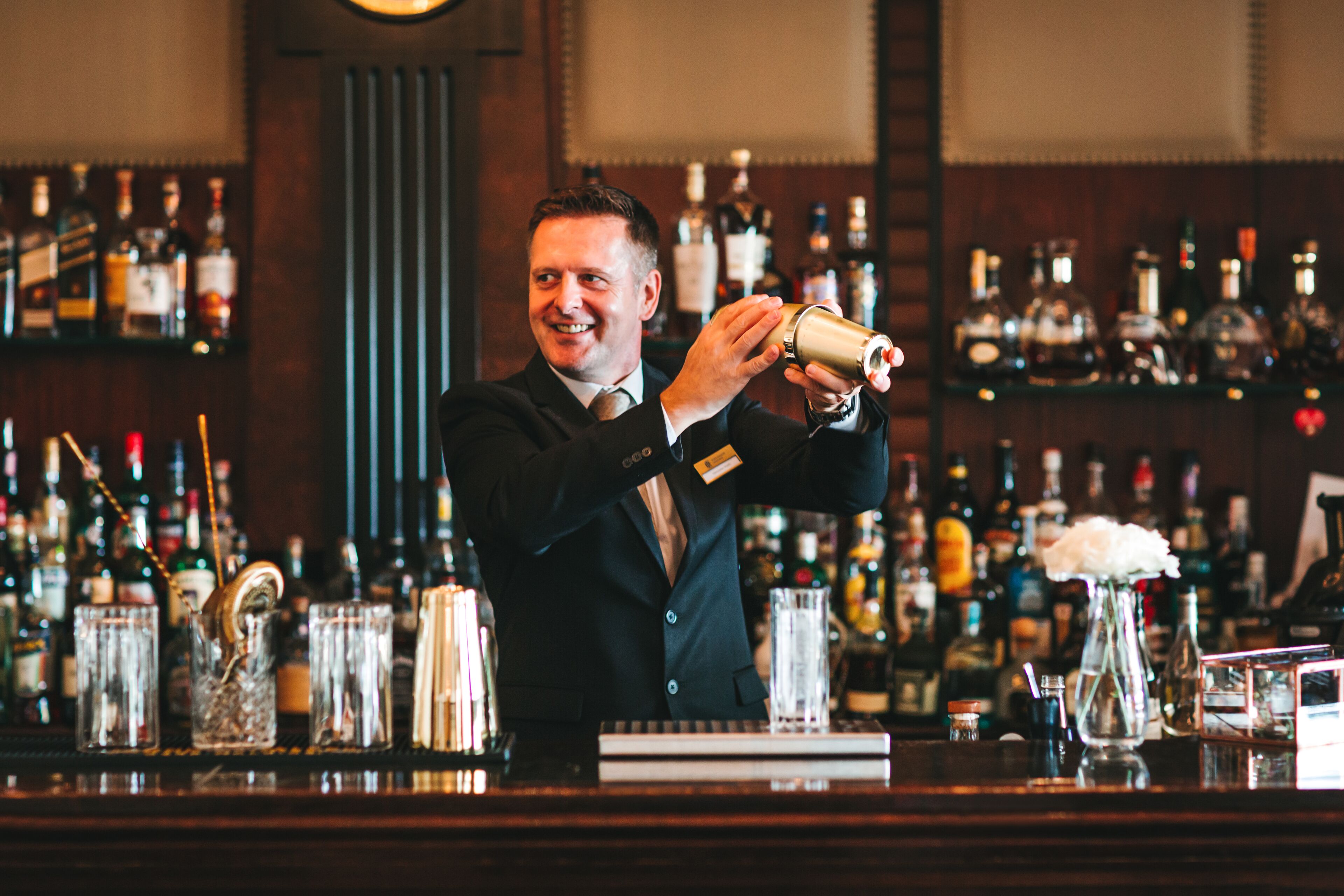 Un barman en train de préparer un cocktail dans un bar, avec des étagères remplies de bouteilles d’alcool en arrière-plan.