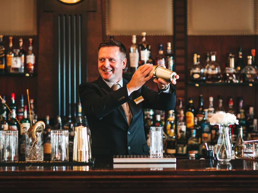 A bartender pouring a drink at a bar with shelves of liquor bottles in the background.