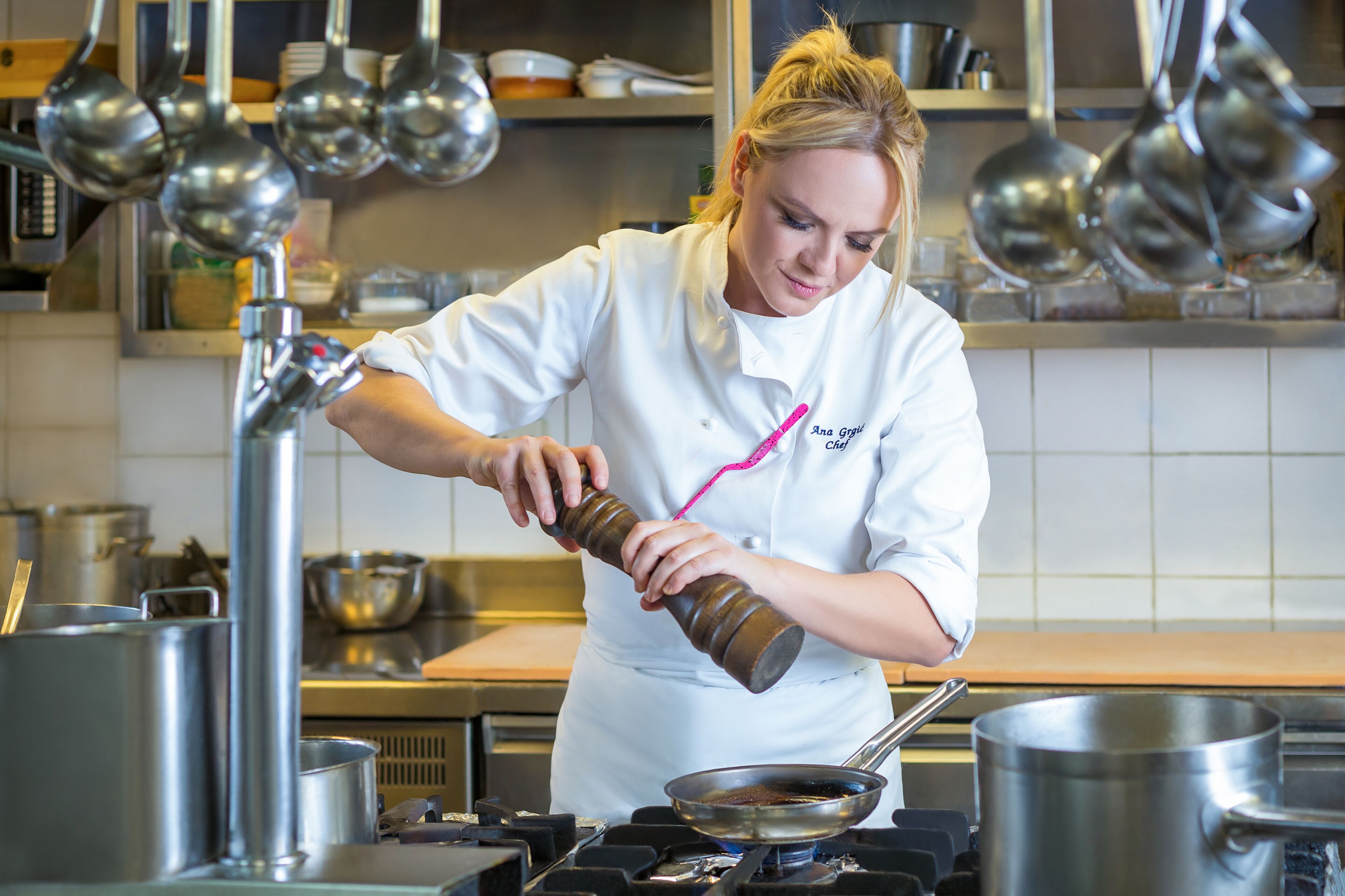 A chef in a white uniform with a pink detail, preparing food in a professional kitchen with stainless steel equipment and pots.