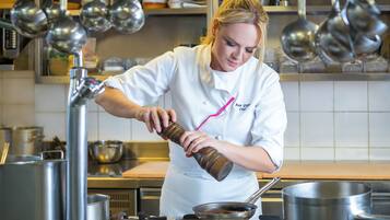 Un chef con uniforme blanco y detalle rosa, preparando comida en una cocina profesional con equipos y ollas de acero inoxidable.