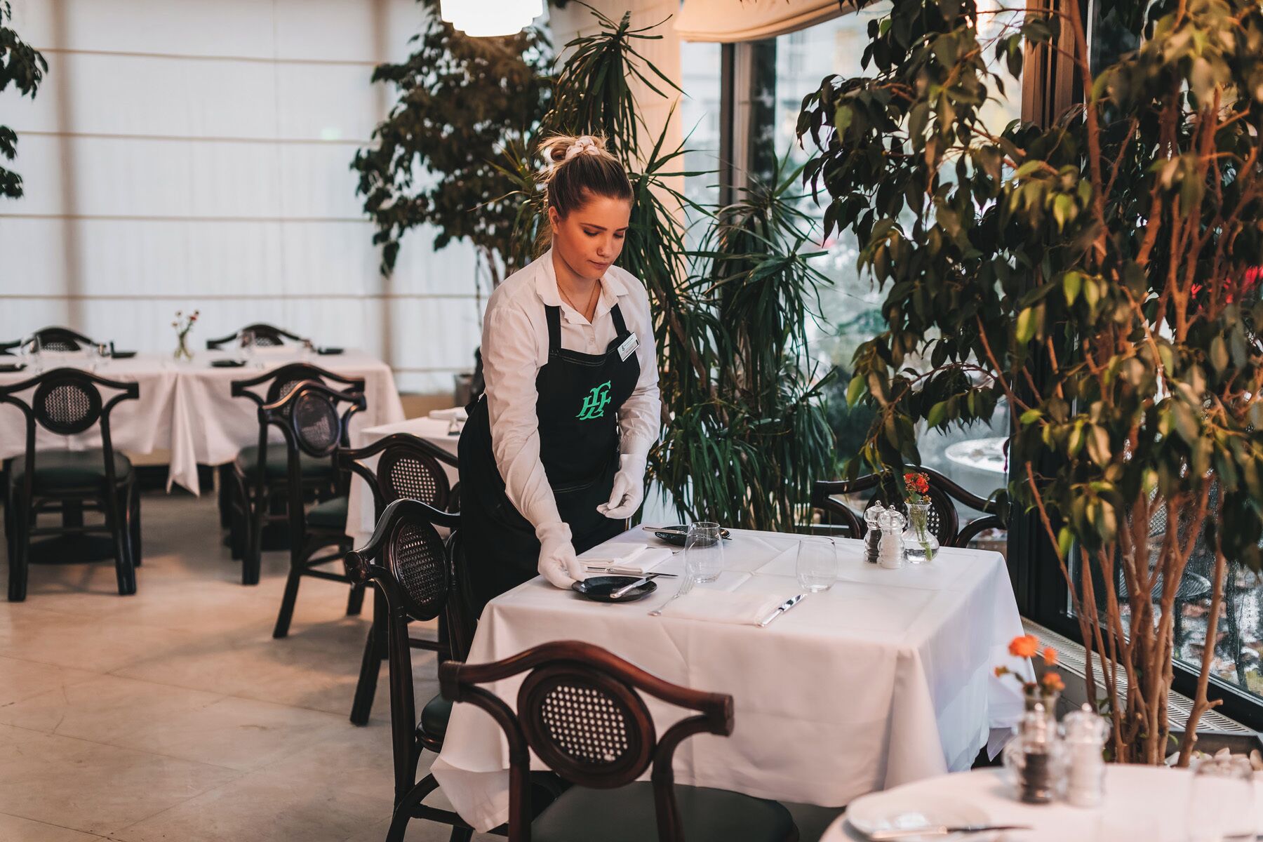 A restaurant with tables set for dining, a waiter attending to a table, and indoor plants.