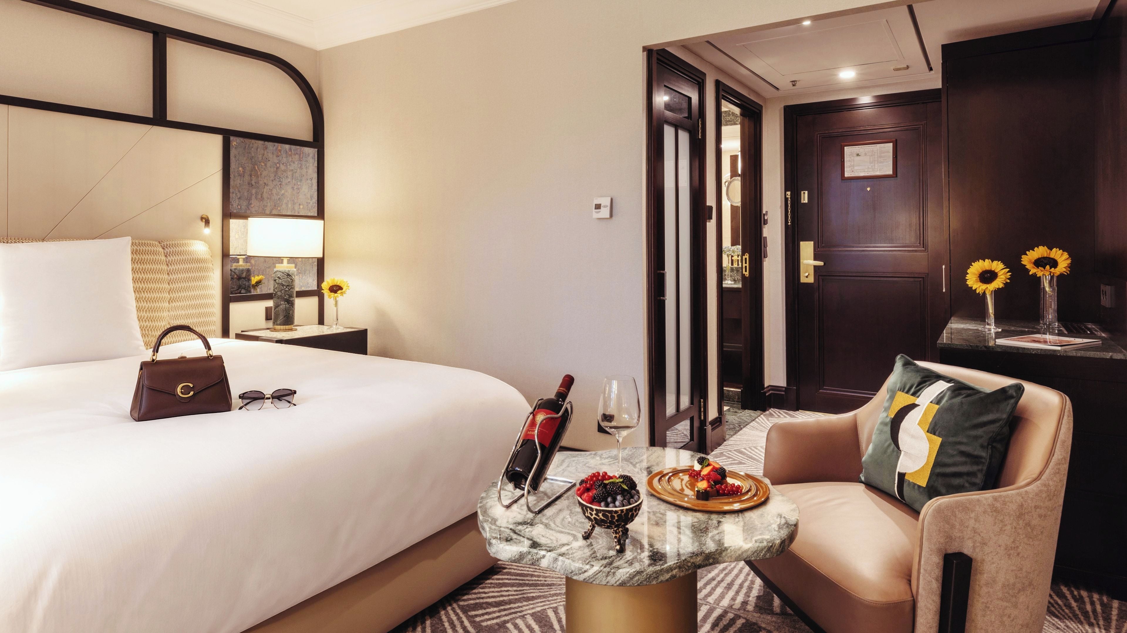 A hotel room with a bed, a sitting area with a table, a minibar, and a door leading to another room.