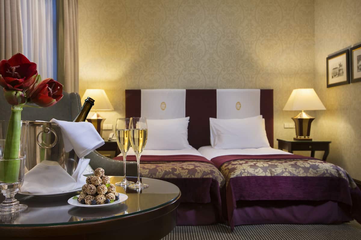 A hotel room with two beds, a glass table with a bottle, glasses, and a plate of snacks.