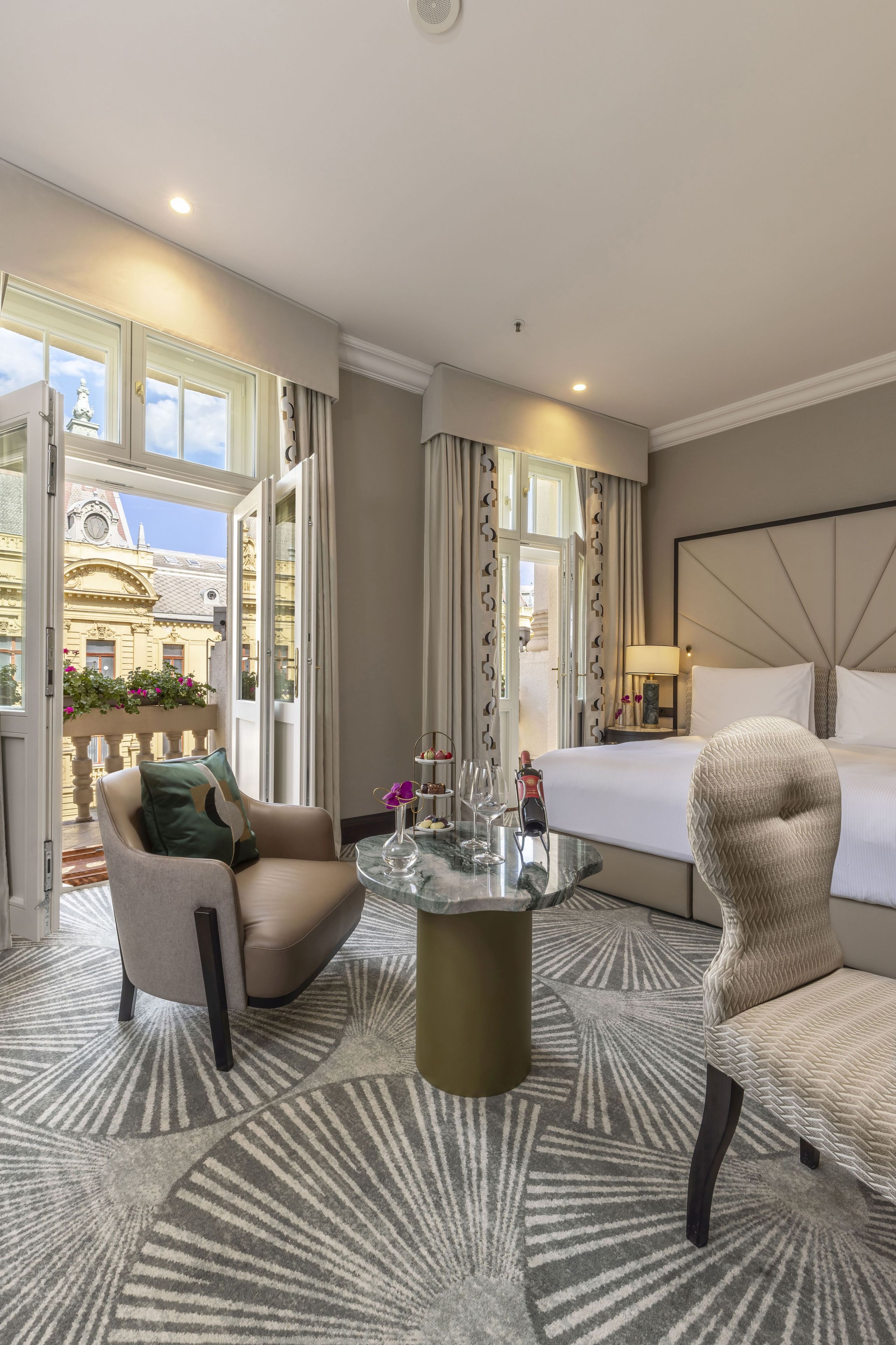 A hotel room with a bed, a sitting area with a chair and a table, a view of the cityscape through large windows, and a balcony with flowers.