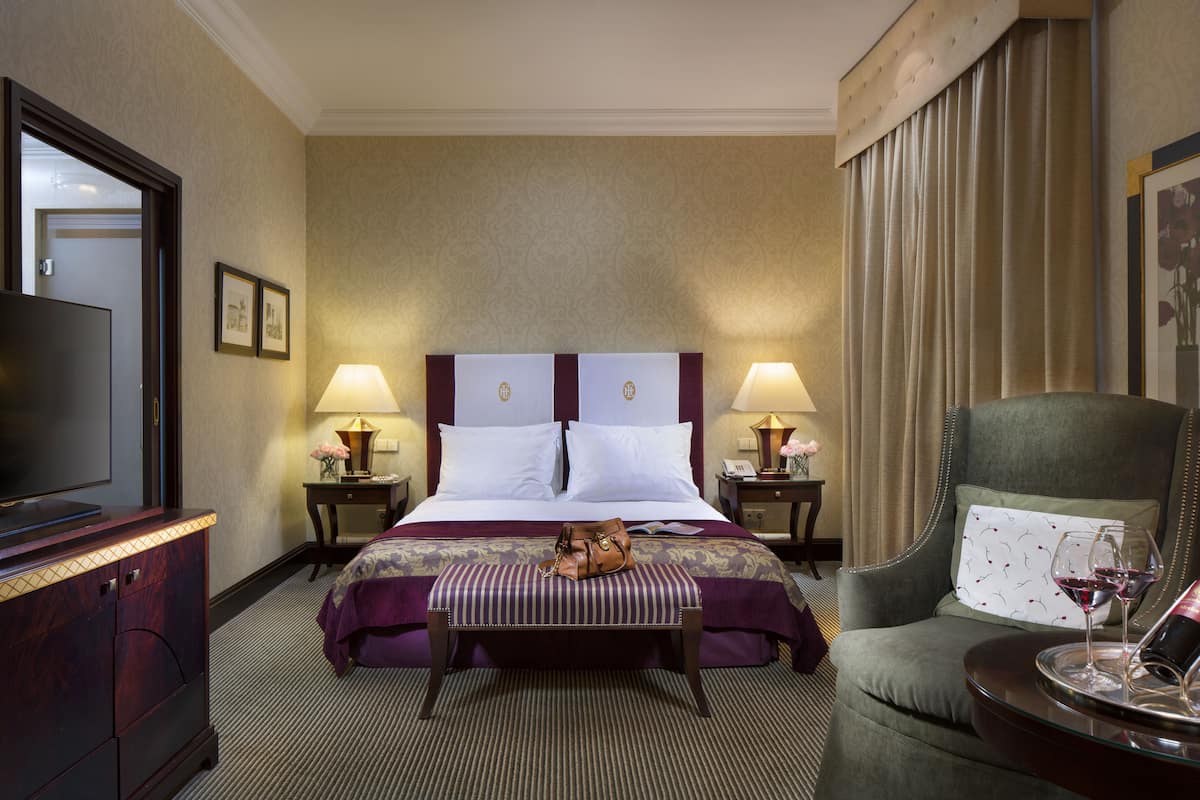 A hotel room with a large bed, two bedside tables with lamps, a television, a green armchair, and a small table with a wine glass.