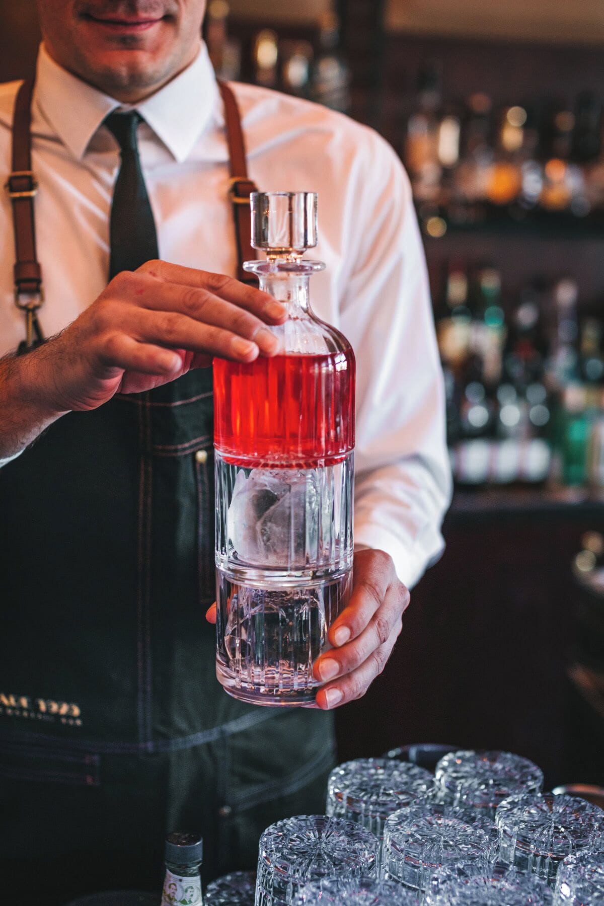 Un barman tenant une grande bouteille de liqueur rouge, avec plusieurs verres disposés au premier plan.