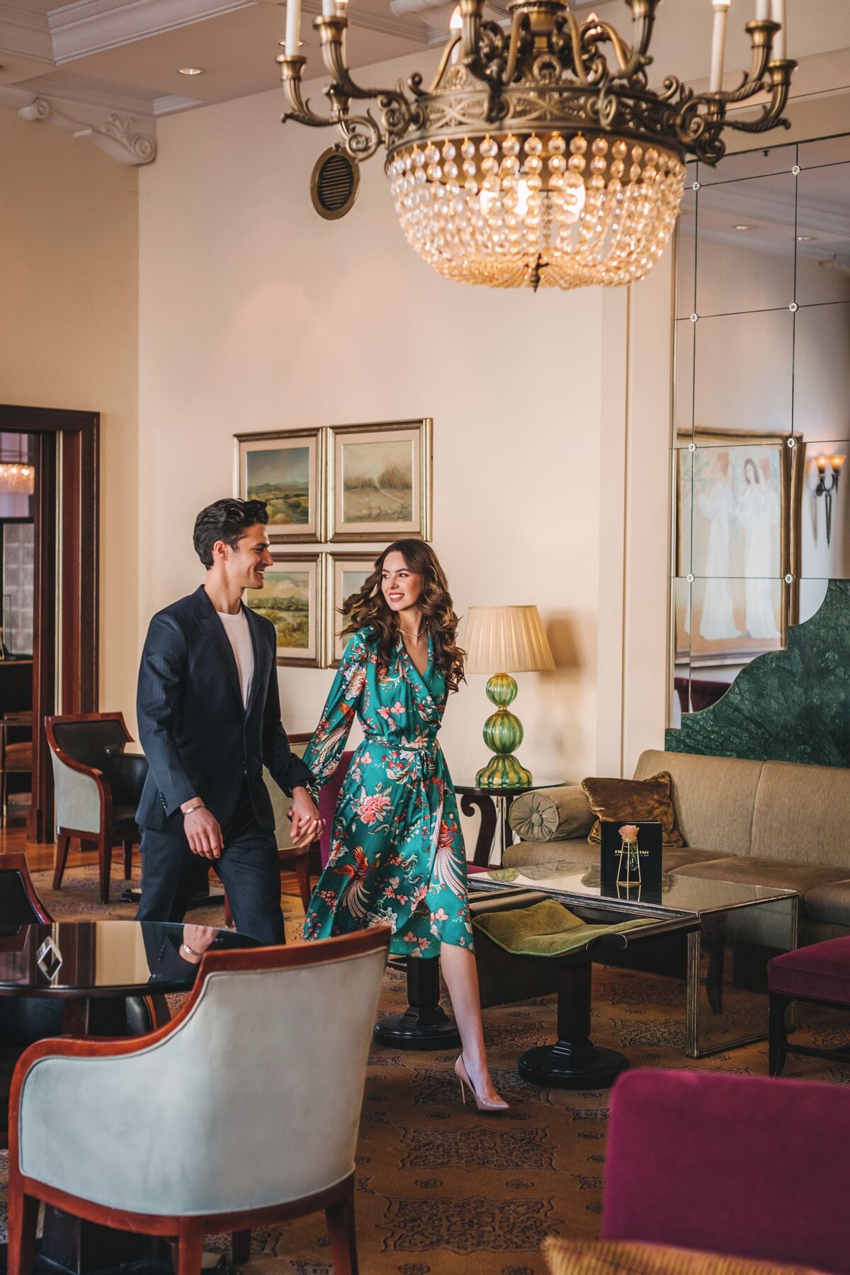 A couple holding hands in a well-lit room with a chandelier, framed artwork on the wall, and a sofa.
