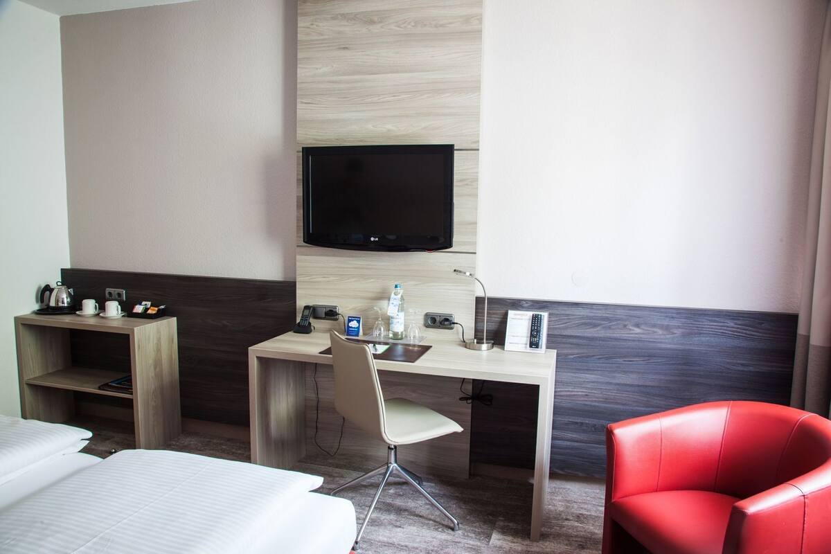 Quarto de hotel com uma cama, uma escrivaninha com cadeira, uma televisão e uma cadeira vermelha.