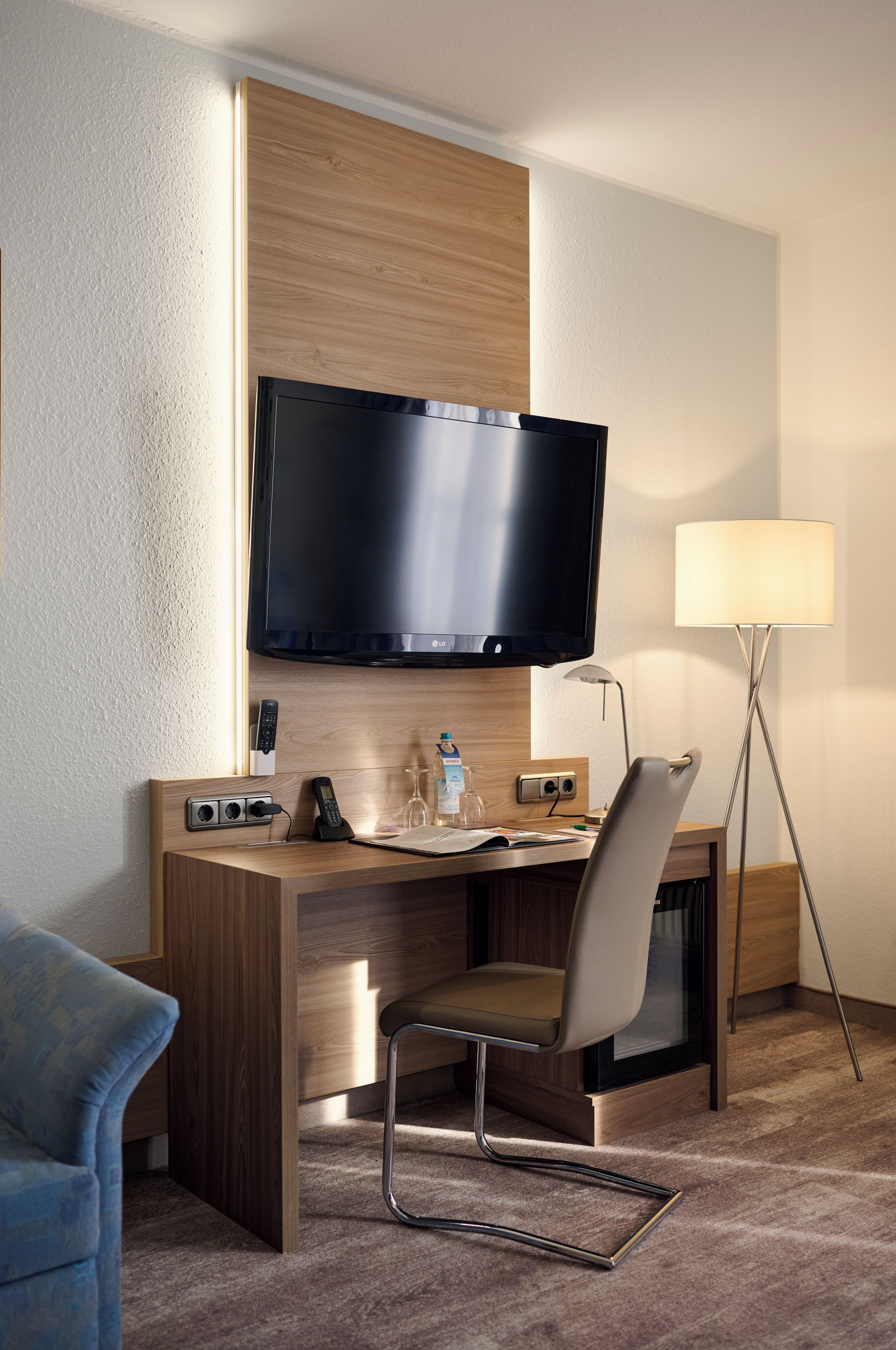 A hotel room with a wooden desk, a flat-screen TV mounted above it, a chair, a desk lamp, and a floor lamp.