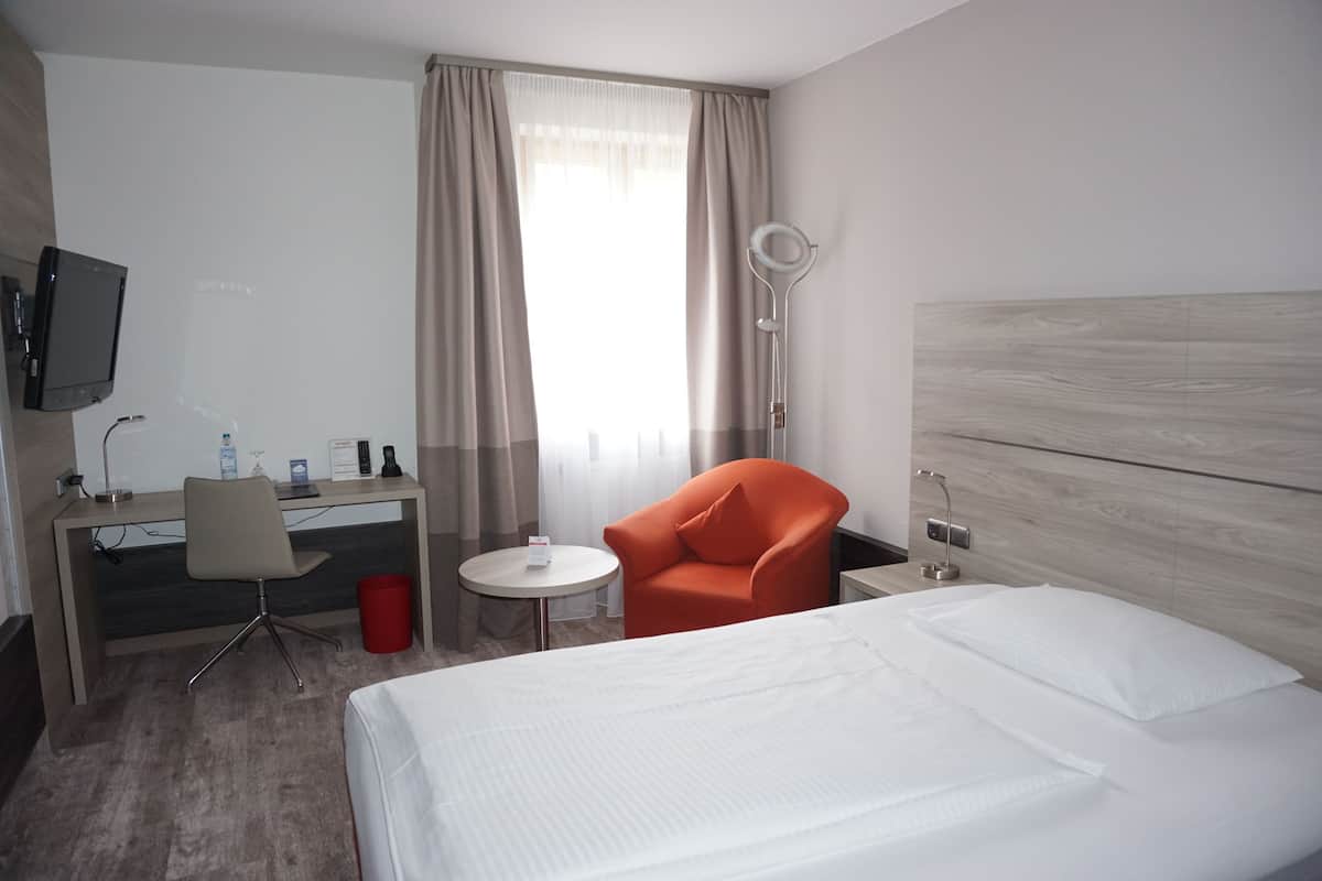 Quarto de hotel com cama, escrivaninha com cadeira, mesinha de centro, lixeira vermelha e TV fixada na parede.