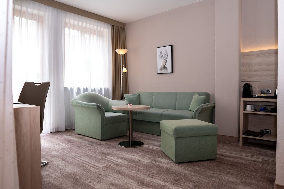Quarto de hotel moderno com sofá de canto, uma pequena mesa redonda, escrivaninha e estante de livros.