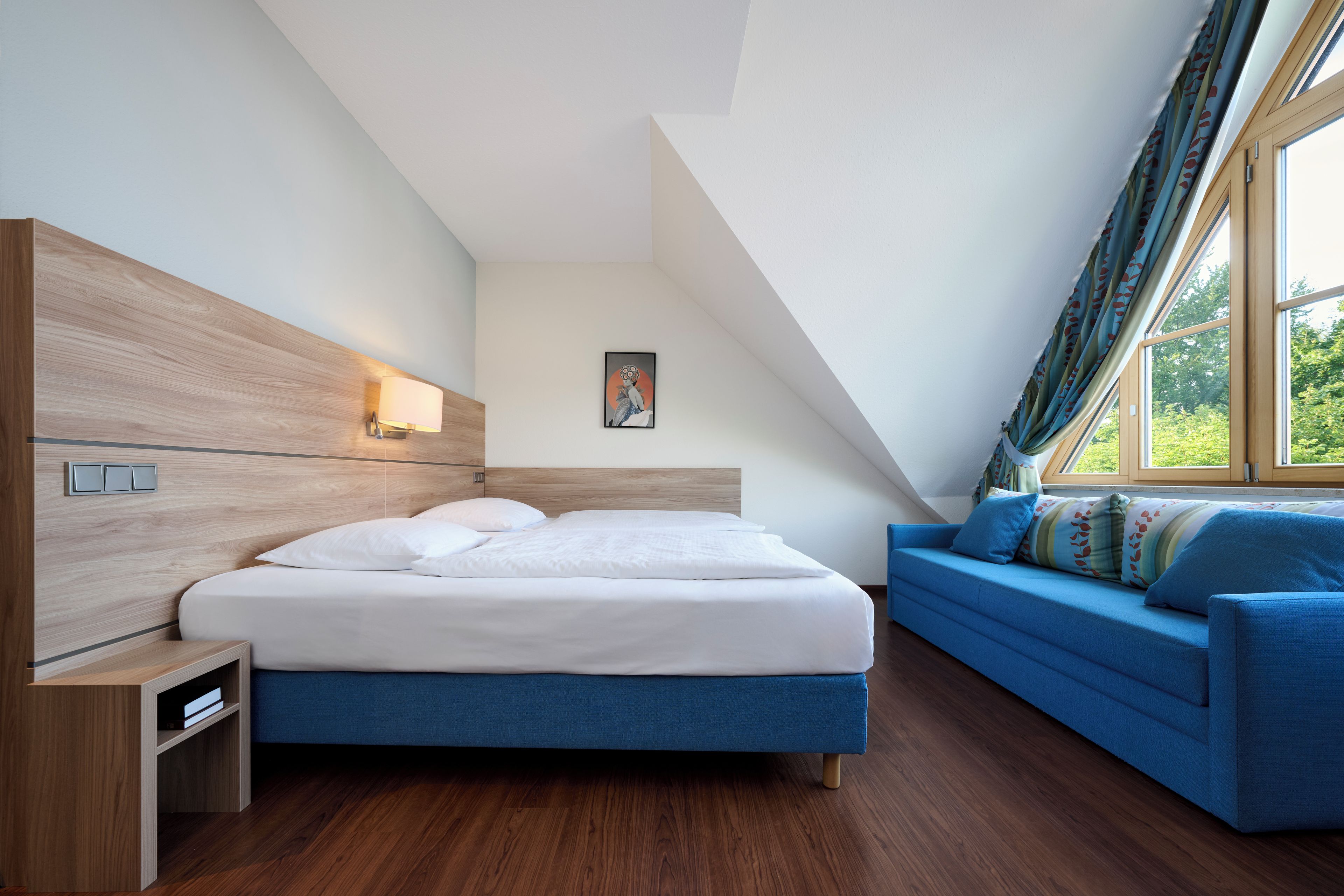 A bedroom with a bed, a blue sofa, a nightstand, a wall-mounted lamp, and a window with curtains.