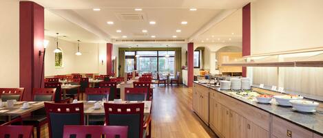 Un restaurante con varias mesas y sillas, una barra de buffet con distintos alimentos, y amplias ventanas que permiten la entrada de luz natural.
