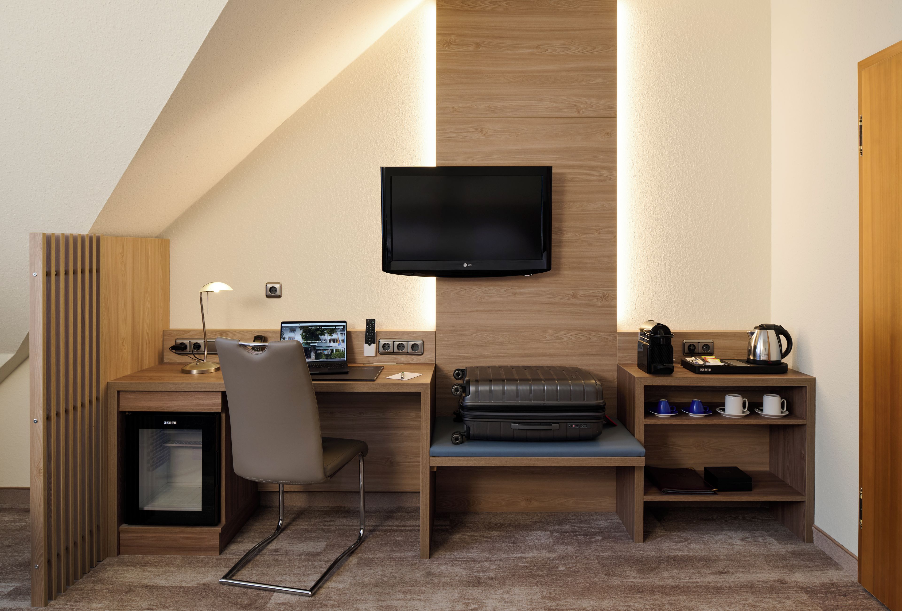 Une chambre d’hôtel de taille réduite, équipée d’un bureau, d’une chaise, d’une télévision et d’un petit coin cuisine.