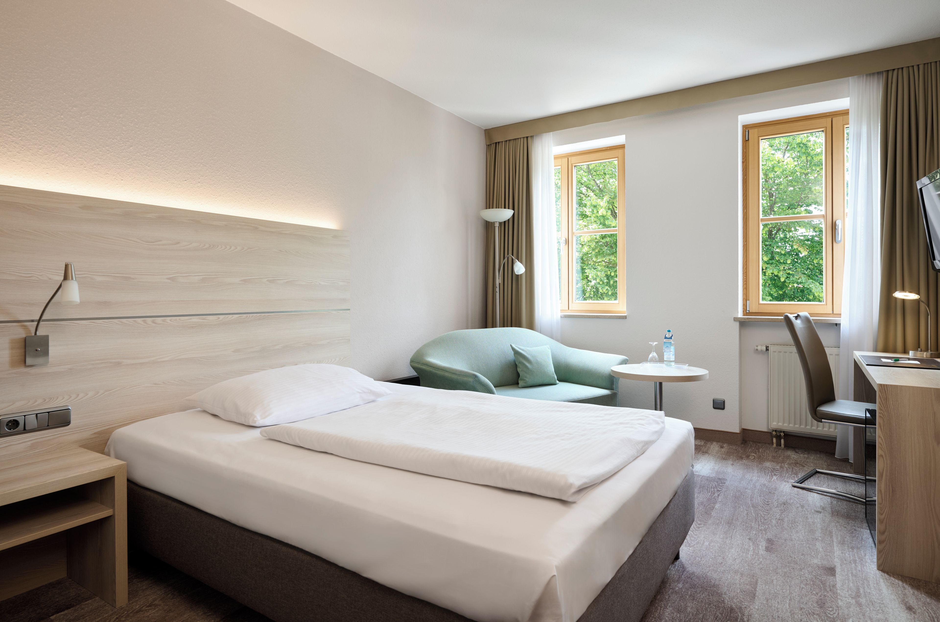 Una habitación de hotel con una cama, un escritorio, una silla, una lámpara y dos ventanas con cortinas.