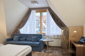 A hotel room with a bed, a blue sofa, a small round table with a lamp, and a view of the outside through a window. - Hotel Kastanienhof (Erding)