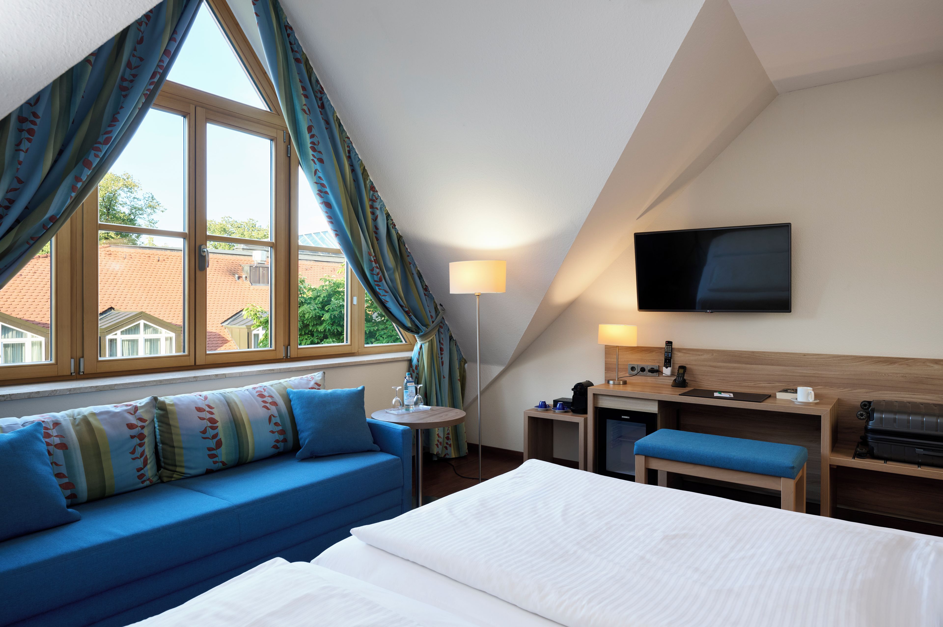Quarto de hotel com cama, sofá, escrivaninha com televisão de tela plana, abajur e vista para o exterior.