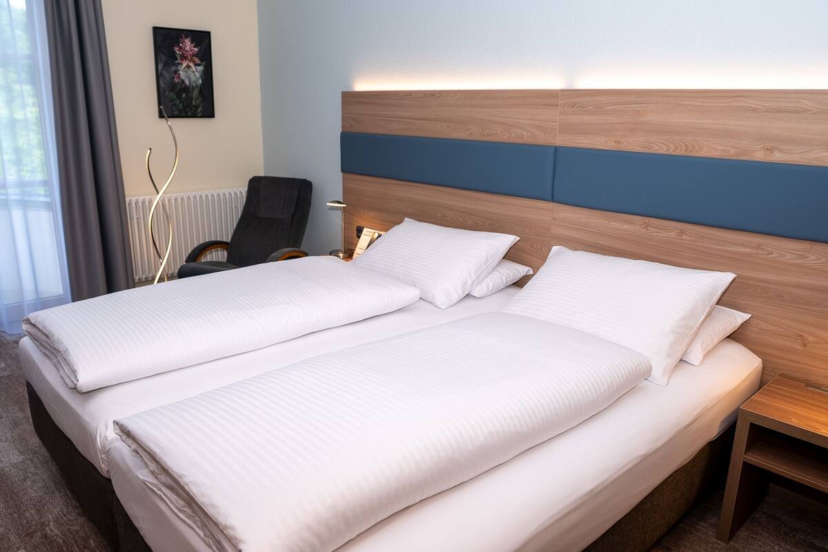 Cama de casal com cabeceira de madeira e detalhe azul, dois travesseiros e mesa de cabeceira com abajur.