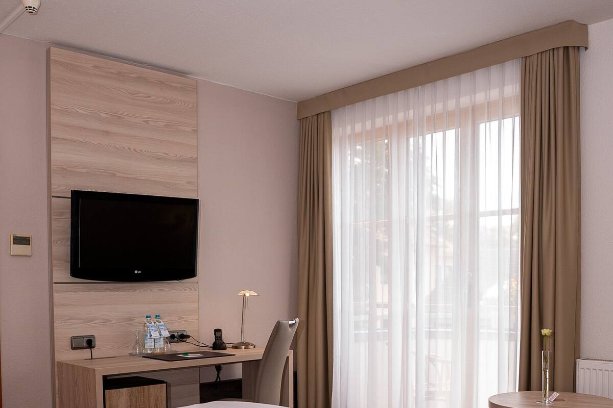 Quarto de hotel com uma cama grande, uma escrivaninha com televisão de tela plana, uma cadeira, uma mesinha de centro com um vaso e uma janela com cortinas translúcidas.