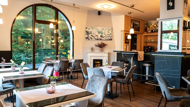 Un restaurant avec des tables rondes, des chaises, une cheminée et de grandes fenêtres donnant sur un espace verdoyant.