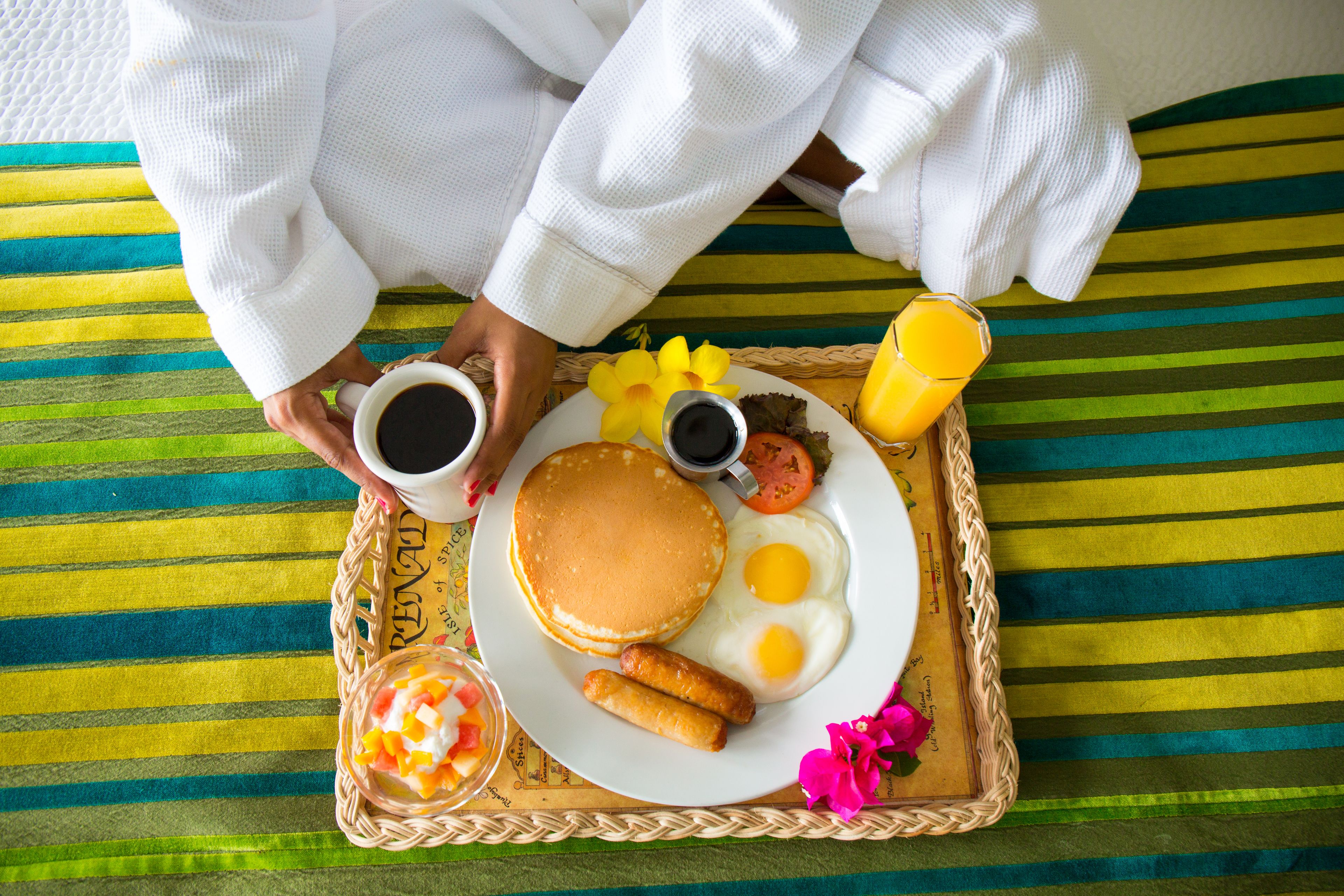 A breakfast tray with pancakes, eggs, sausages, a cup of coffee, and a glass of orange juice on a striped bedspread.