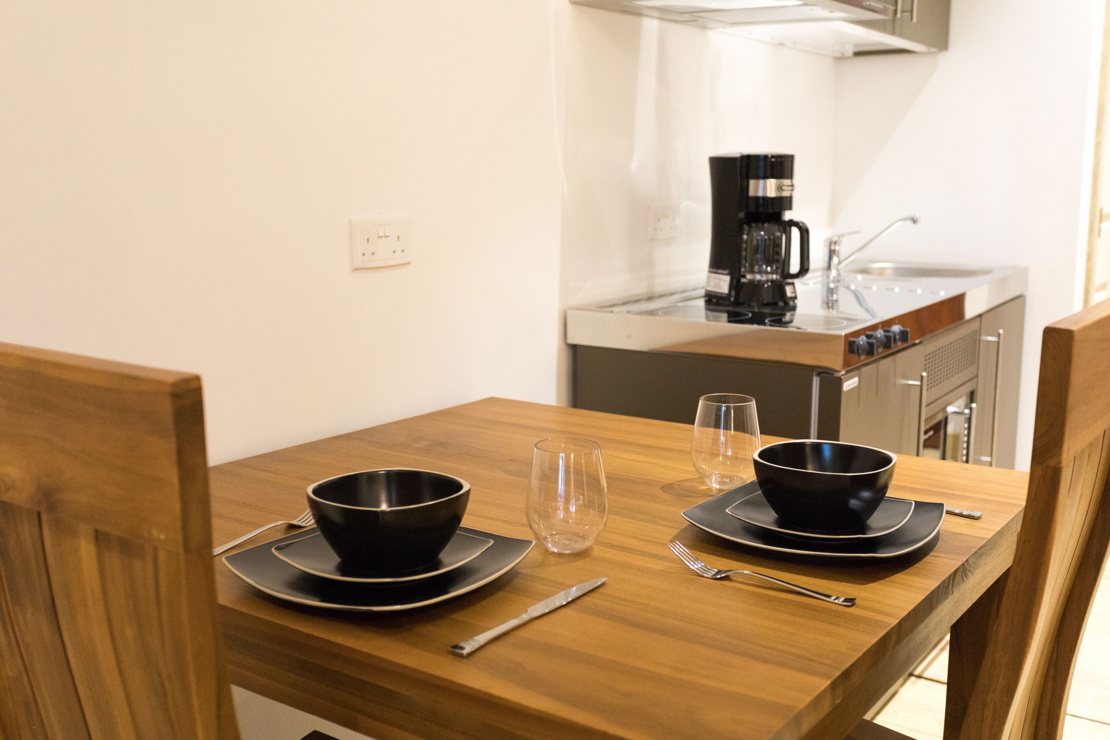 Una mesa de comedor con dos cuencos negros, dos copas de vino, una cafetera y un cuchillo sobre una mesa de madera en una cocina.