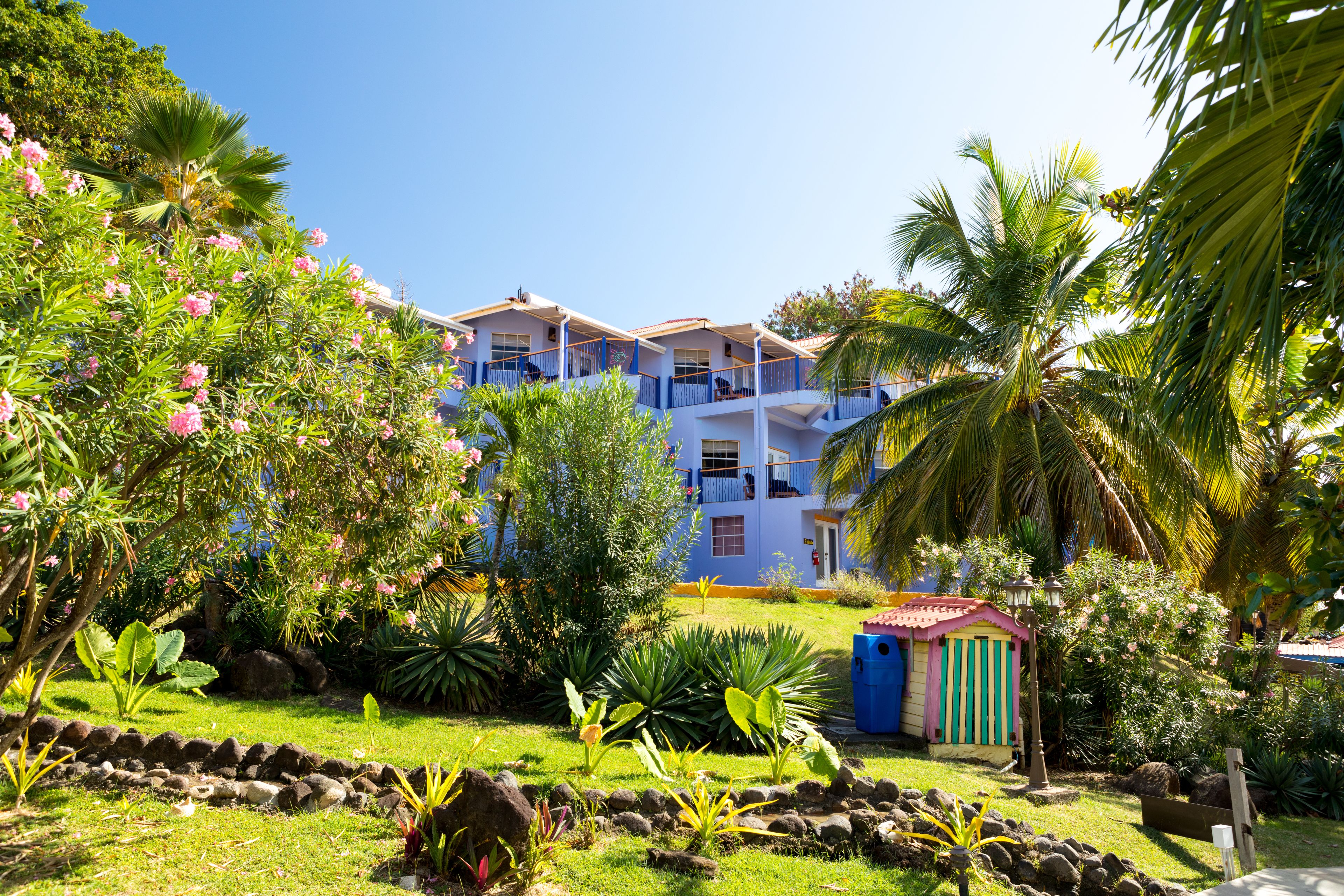 Un hotel con varios balcones, exuberantes jardines tropicales y una pequeña casita de juegos.
