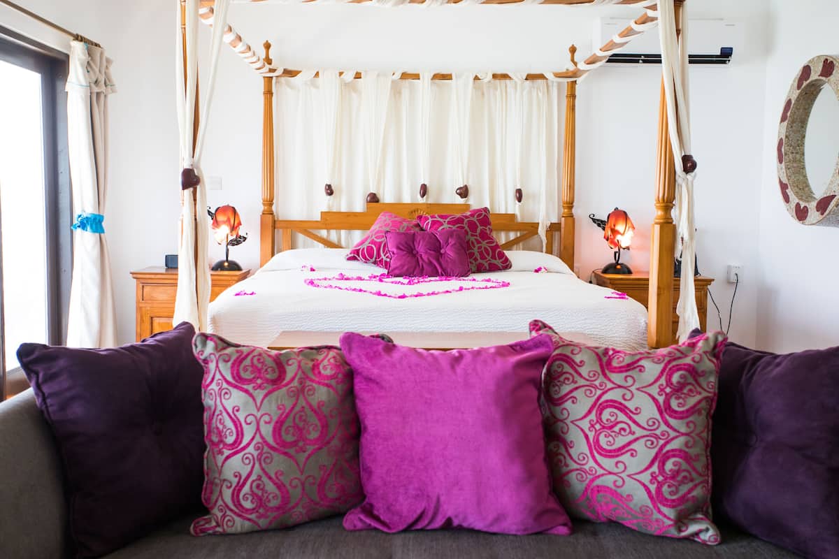 A four-poster canopy bed with a white bedspread and multiple pillows, including a prominent purple one.