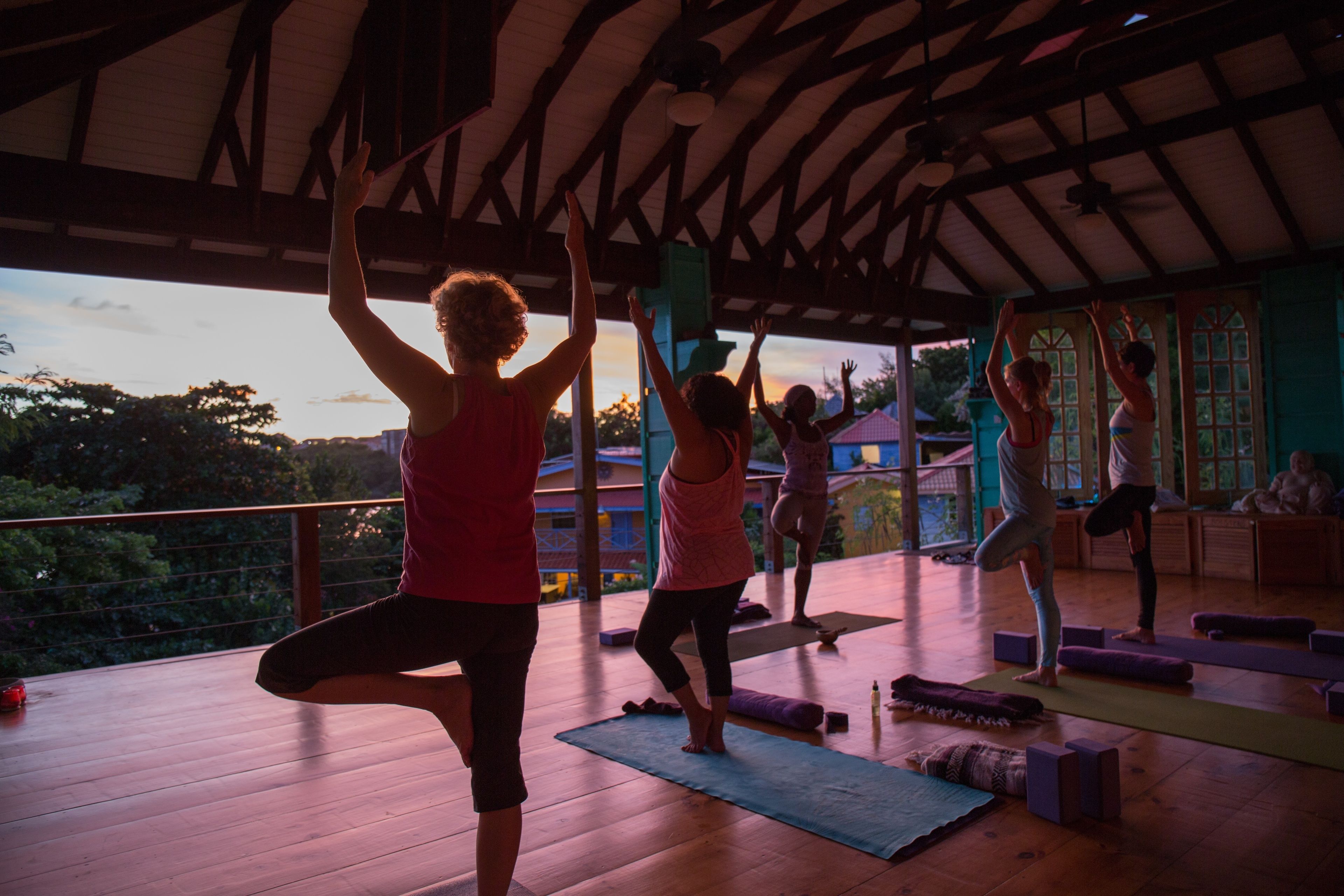 Un cours de yoga dans une pièce au sol en bois, avec vue sur des arbres et un bâtiment en arrière-plan.