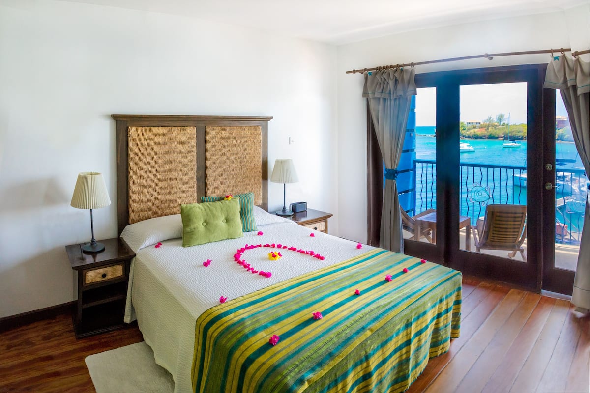 A hotel room with a bed, a nightstand, a lamp, a window with curtains, and a view of the sea.