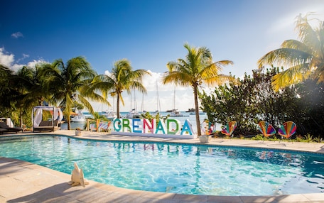 A swimming pool with a sign reading "Grenada," surrounded by palm trees and lounge chairs.