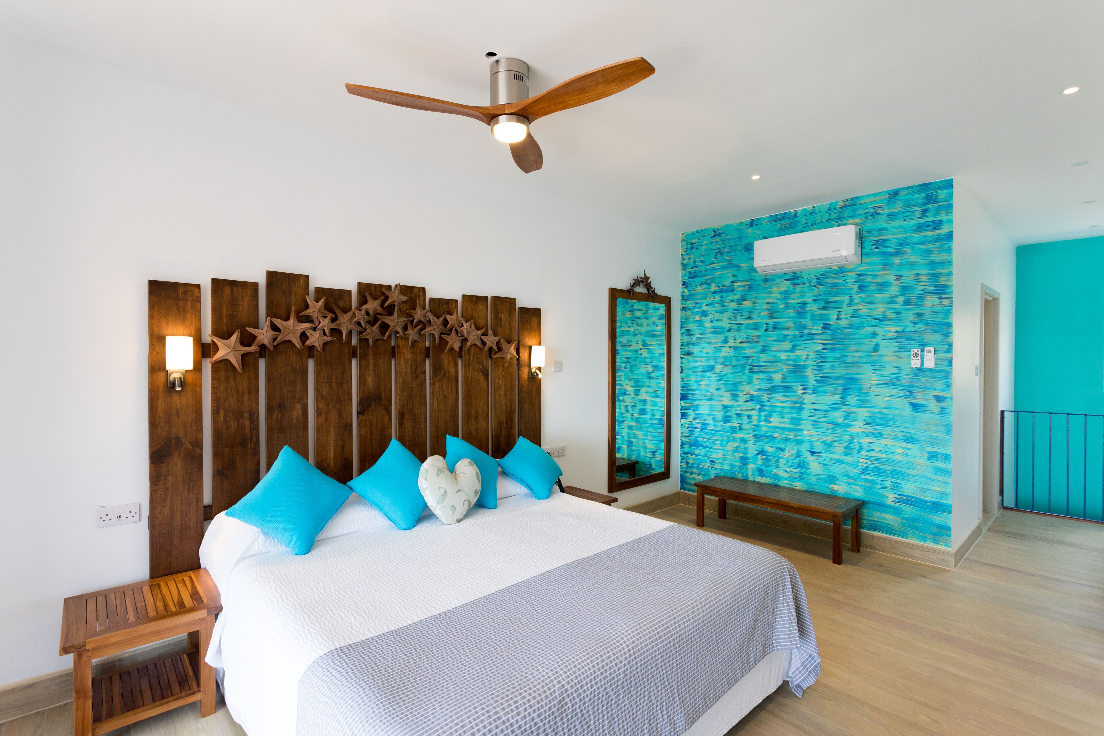 A bedroom with a wooden headboard, a ceiling fan, a large bed with blue and white pillows, a wooden bench, and a blue textured wall.