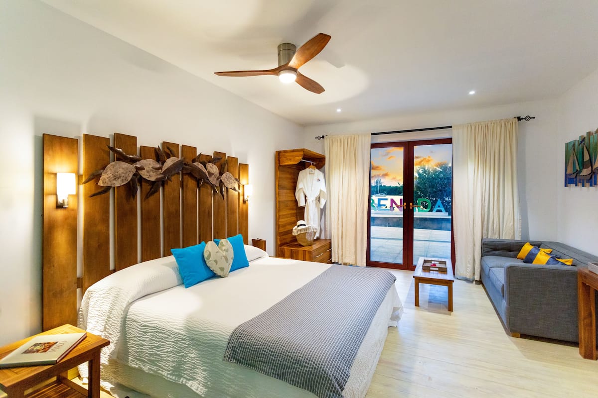 A hotel room with a large bed, a wooden headboard, a ceiling fan, a wooden nightstand, a grey sofa, and a view of a building through the glass door.