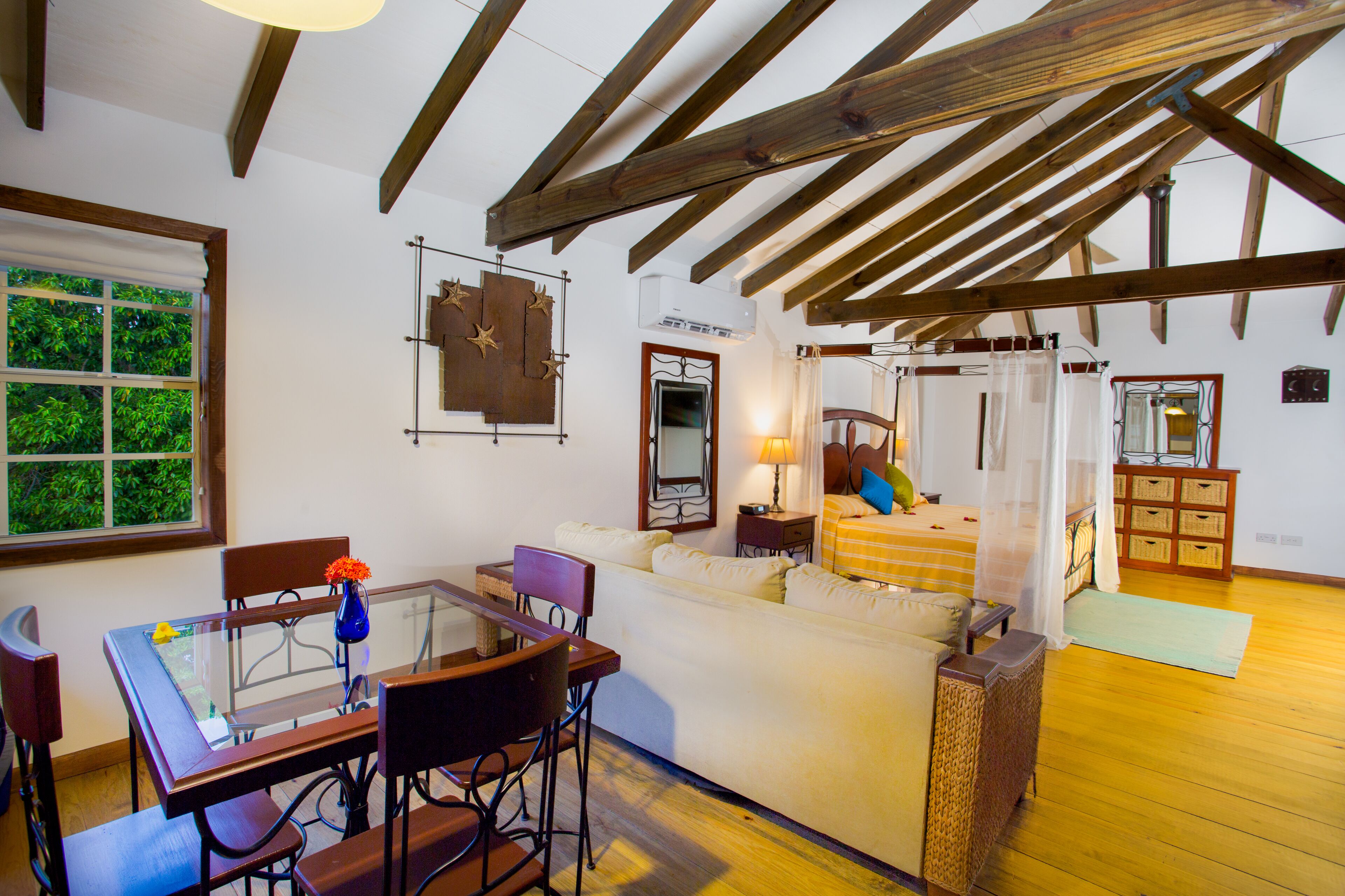 A room with a wooden ceiling, a glass dining table, a sofa, and a bed with a canopy.