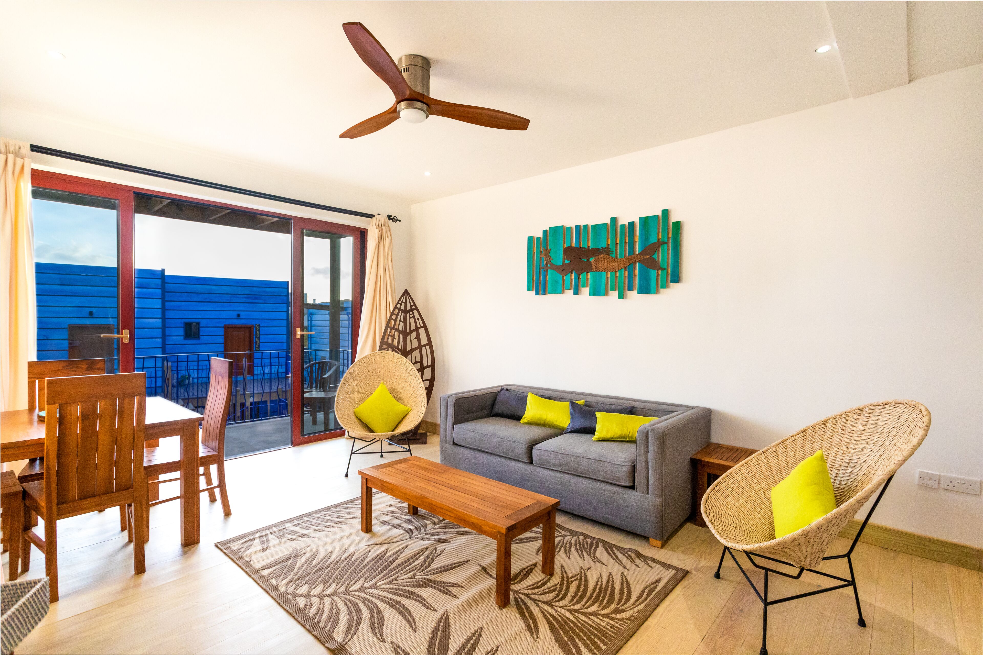 A living room with a sofa, chairs, a coffee table, a ceiling fan, and a large window with a view of a blue building.