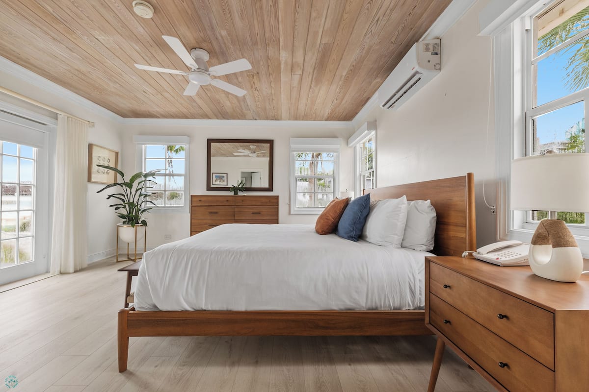 A bedroom with a wooden ceiling, a bed with white bedding, a bedside table with a lamp, and a window with a view of palm trees.