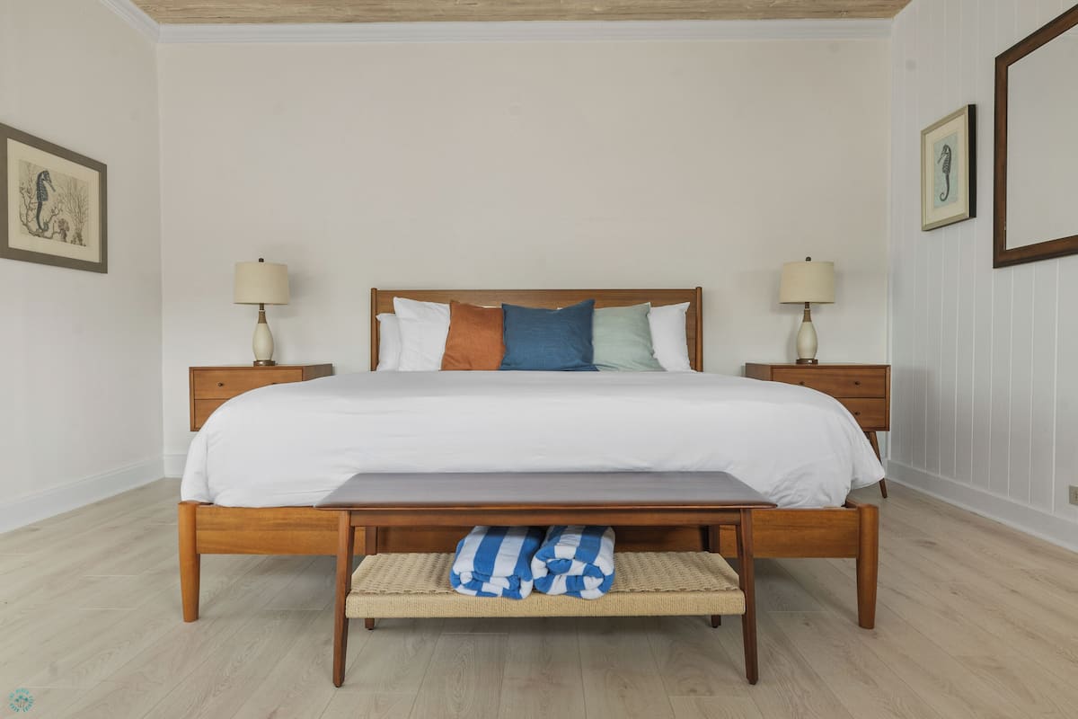 A neatly made bed with a wooden frame, white bedding, and colorful pillows. There are two bedside tables with lamps, a wooden bench at the foot of the bed, and a framed picture on the wall.