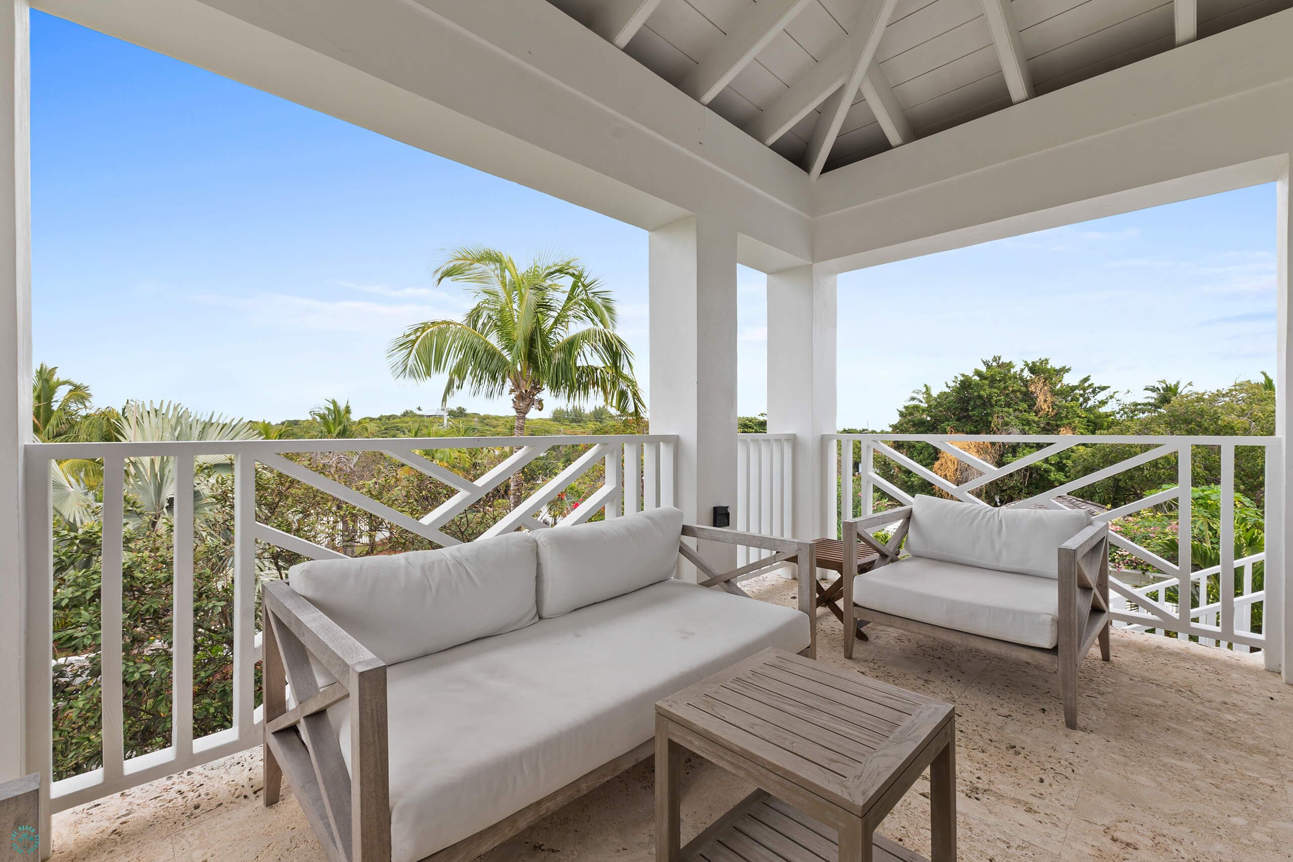 A covered outdoor seating area with white railings, a wooden table, and a white sofa with cushions.