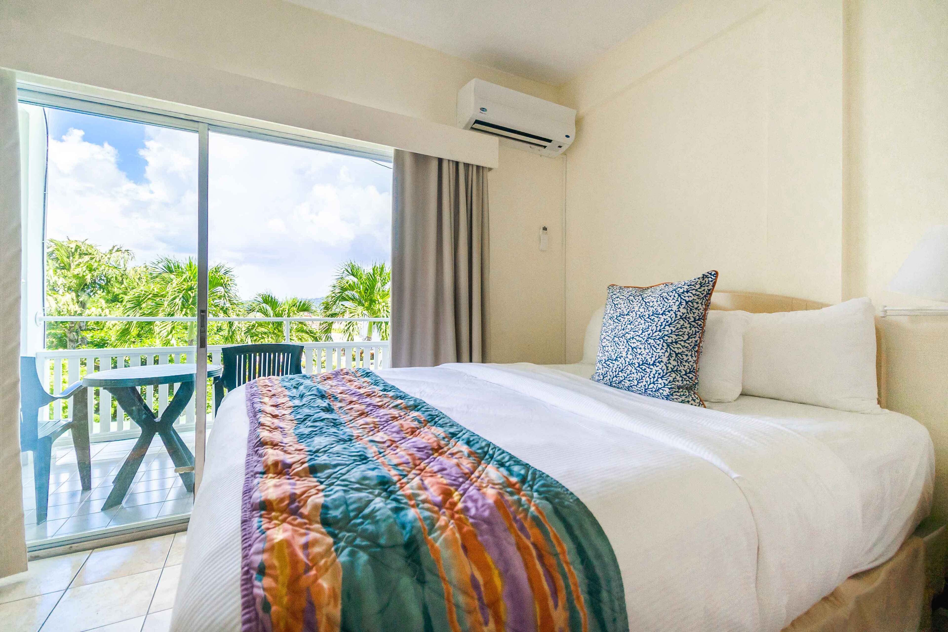 A hotel room with a large bed, a balcony with a view of palm trees, and an air conditioning unit on the wall.