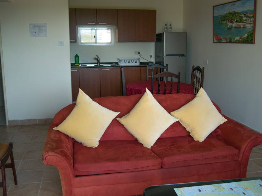 A red sofa with three pillows, a wooden chair, and a kitchen area with wooden cabinets and a refrigerator.