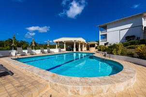 A swimming pool with lounge chairs and a covered area, surrounded by a white building and greenery.