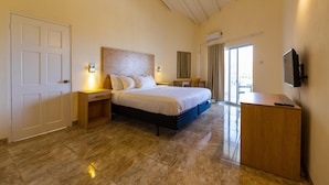 A bedroom with a bed, bedside tables, a desk, a television, and a sliding glass door to a balcony. - Siesta Hotel (St. George's)