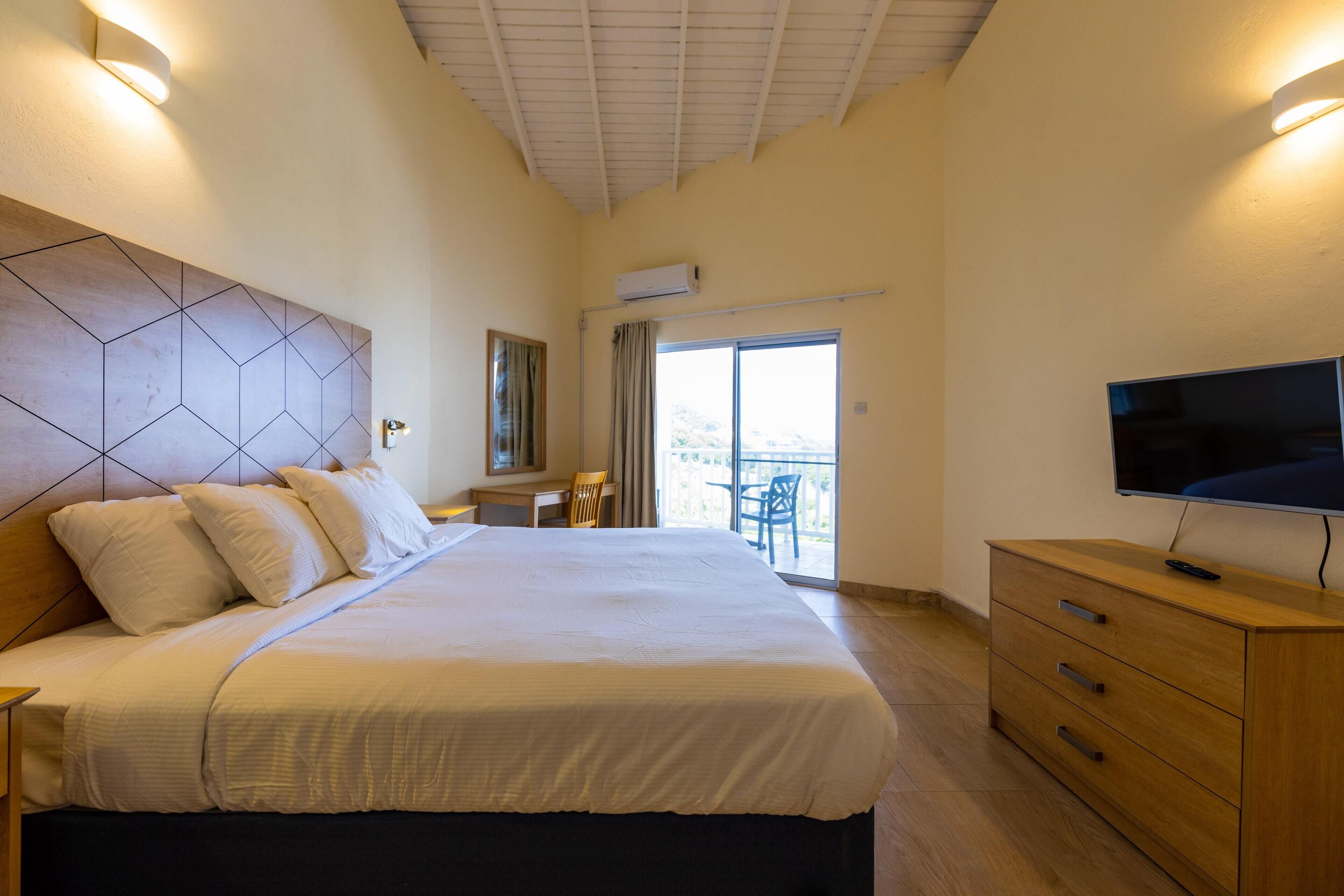 A hotel room with a large bed, a TV, a wooden dresser, a desk, and a balcony with a view.