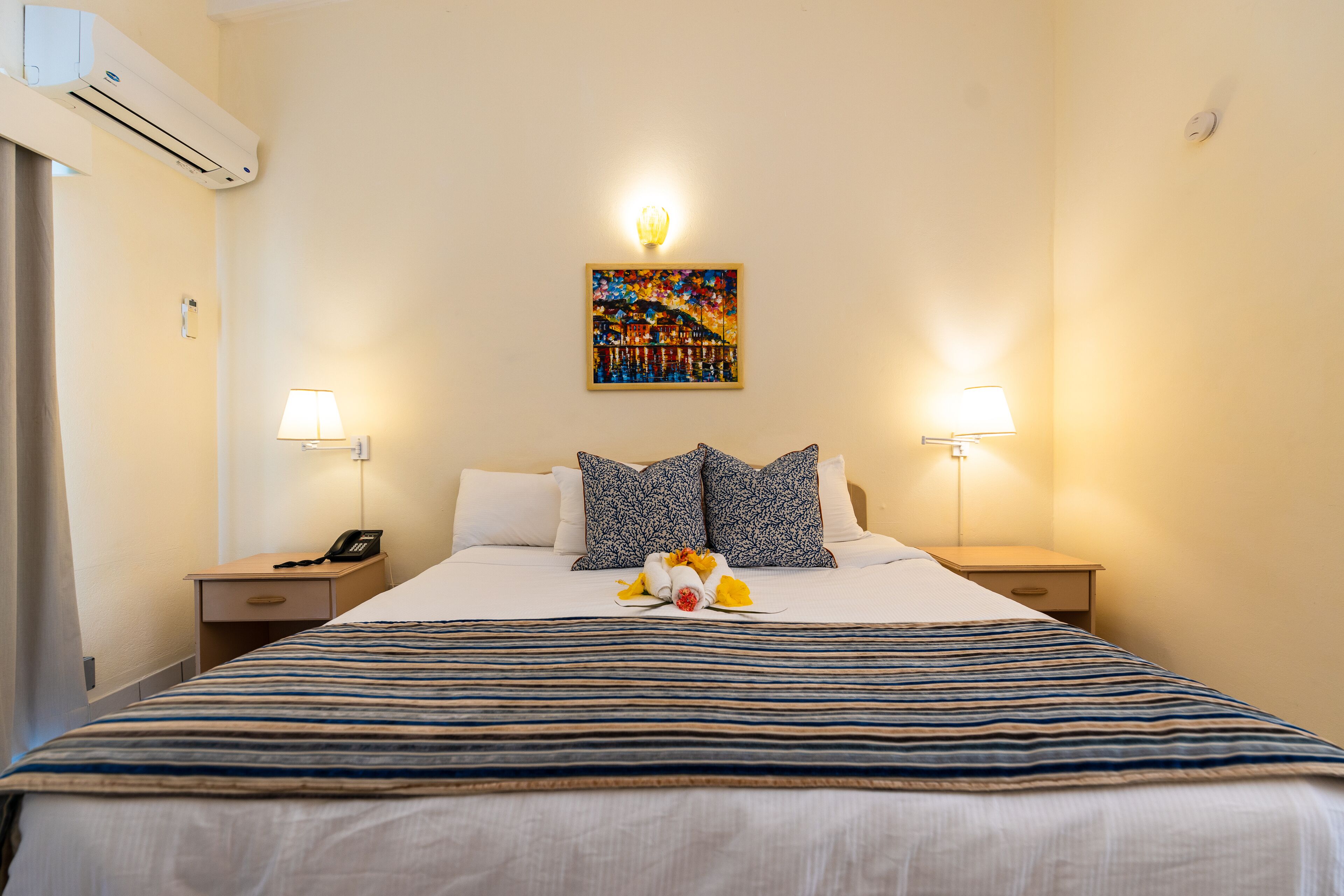 A hotel room with a large bed, two bedside tables with lamps, a wall-mounted air conditioner, and a painting above the bed.