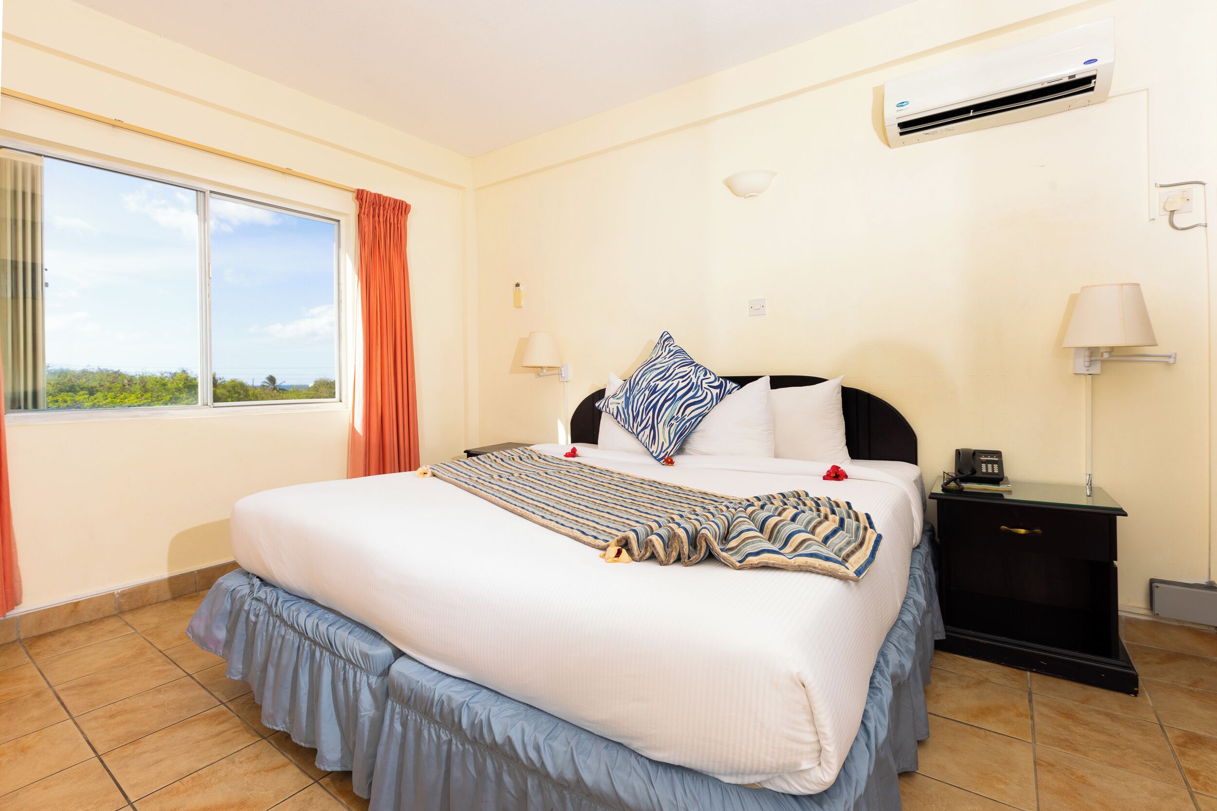 Habitación de hotel con una cama grande, una mesita de noche, una lámpara, aire acondicionado y vistas al exterior a través de una ventana.