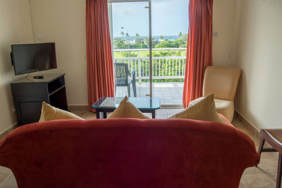 A room with a red sofa, a TV, a small table, and a balcony with a view of the sea.